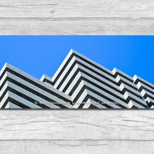 May include: Architectural photograph of a building with a geometric design. The building has a series of gray, horizontal, stepped layers against a bright blue sky. The image is a long, horizontal rectangle.