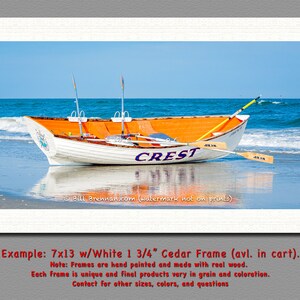 May include: A framed print of a white and orange rowboat on a beach, with the word "CREST" on the side. The boat has two oars and is set against a blue sky and ocean. The frame is white.