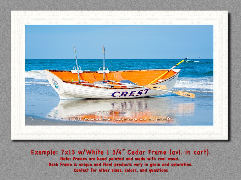 May include: A framed print of a white and orange rowboat on a beach, with the word "CREST" on the side. The boat has two oars and is set against a blue sky and ocean. The frame is white.