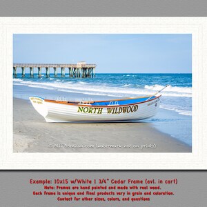 May include: A framed print showing a white boat with a blue stripe on a sandy beach, with the words "NORTH WILDWOOD" on the side. The sea and a pier are visible in the background. The image is framed in a white wooden frame.