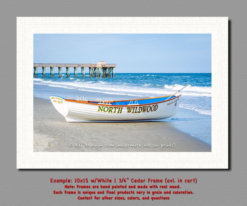 May include: A framed print showing a white boat with a blue stripe on a sandy beach, with the words "NORTH WILDWOOD" on the side. The sea and a pier are visible in the background. The image is framed in a white wooden frame.
