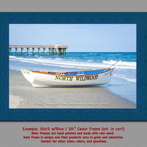 May include: A framed print showing a white boat on a sandy beach with the text "NORTH WILDWOOD". The boat has a blue stripe and is near the sea with a pier in the background. The frame is blue.