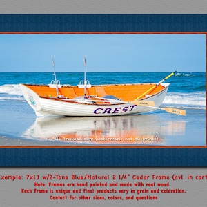 May include: A framed print featuring a white and orange boat on a beach with the word "CREST" on its side. The boat has oars and is set against a blue ocean and sky. The frame is two-tone blue and natural.