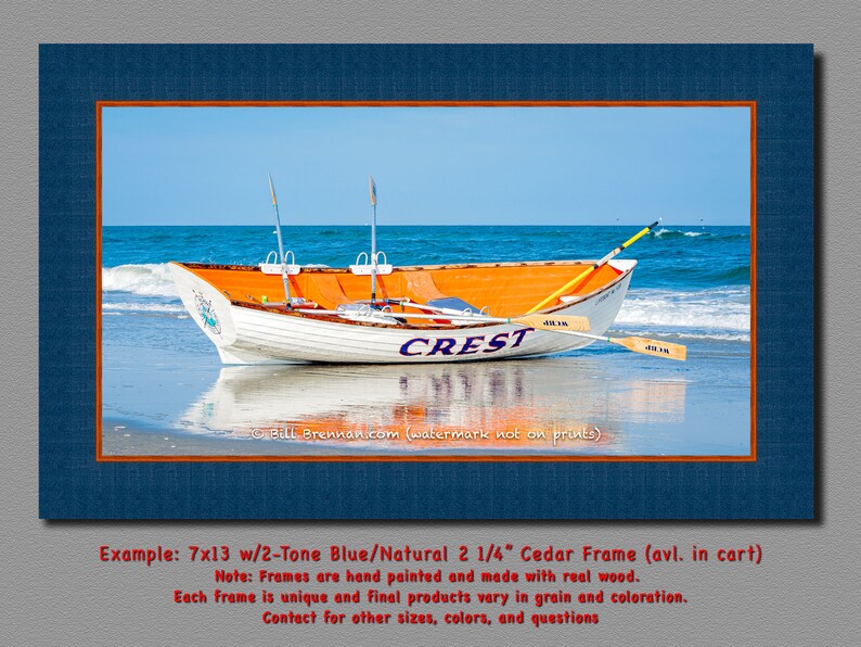 May include: A framed print featuring a white and orange boat on a beach with the word "CREST" on its side. The boat has oars and is set against a blue ocean and sky. The frame is two-tone blue and natural.
