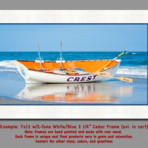May include: A framed print featuring a white and orange rowboat named "CREST" on a sandy beach with blue water and sky. The boat has oars and is framed in a white wooden frame with a blue border. The image is a landscape scene.