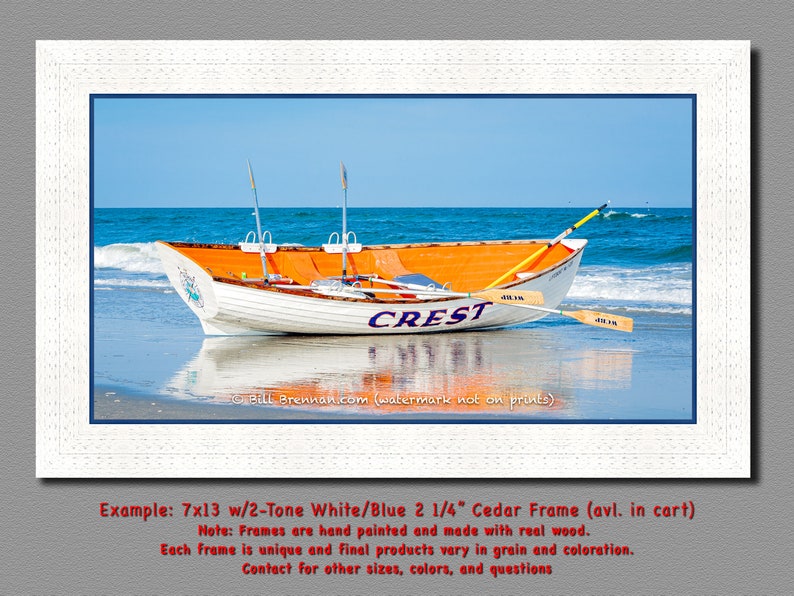 May include: A framed print featuring a white and orange rowboat named "CREST" on a sandy beach with blue water and sky. The boat has oars and is framed in a white wooden frame with a blue border. The image is a landscape scene.