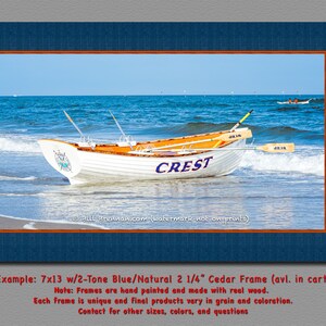 K&ouml;nnte beinhalten: Gerahmter Druck mit einem wei&szlig;en Ruderboot namens "CREST" am Strand mit blauem Wasser und Himmel. Das Boot hat orangefarbene Akzente und Ruder. Der Rahmen ist zweifarbig blau und Naturholz. Die Druckgr&ouml;&szlig;e betr&auml;gt 17,78cm x 33,02cm.