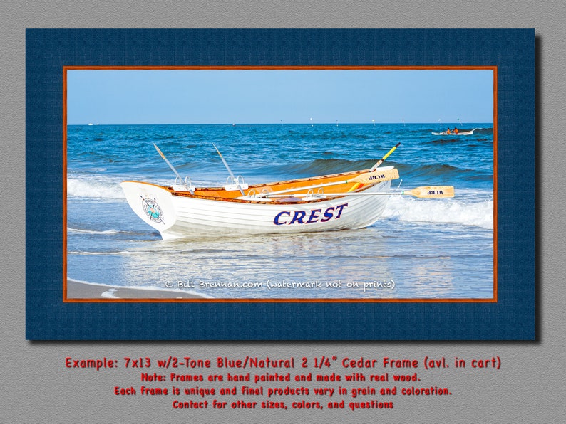 K&ouml;nnte beinhalten: Gerahmter Druck mit einem wei&szlig;en Ruderboot namens "CREST" am Strand mit blauem Wasser und Himmel. Das Boot hat orangefarbene Akzente und Ruder. Der Rahmen ist zweifarbig blau und Naturholz. Die Druckgr&ouml;&szlig;e betr&auml;gt 17,78cm x 33,02cm.