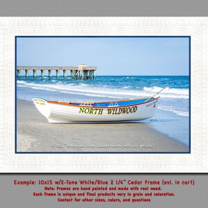 May include: A framed print showing a white boat with blue detailing on a sandy beach. The boat is inscribed with "NORTH WILDWOOD" in green. A wooden pier stretches into the blue sea under a clear sky. The frame is white.