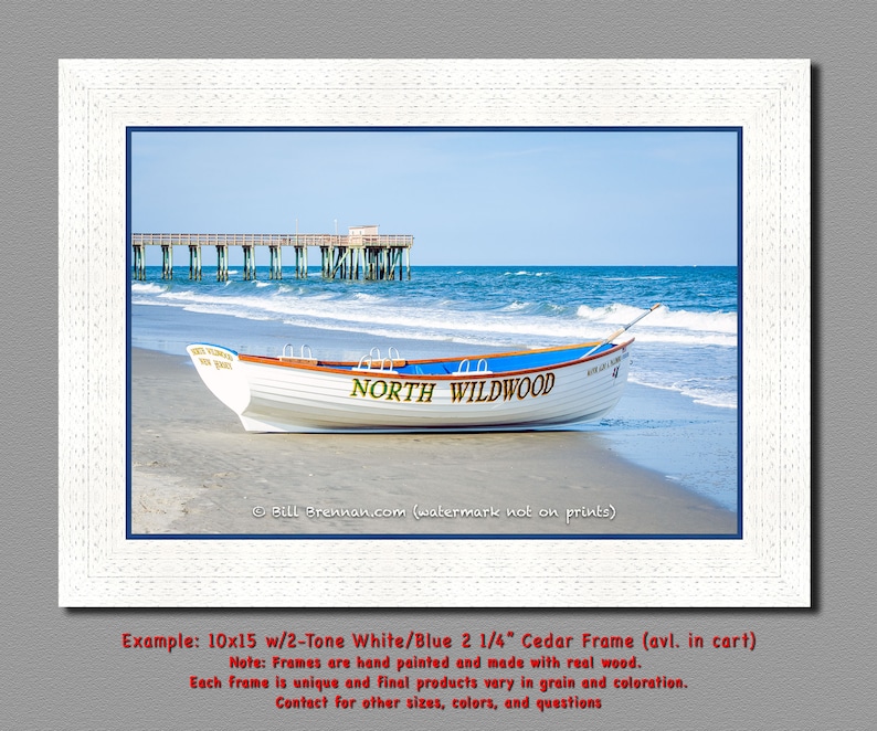 May include: A framed print showing a white boat with blue detailing on a sandy beach. The boat is inscribed with "NORTH WILDWOOD" in green. A wooden pier stretches into the blue sea under a clear sky. The frame is white.