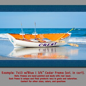 May include: A framed print of a white and orange boat on a beach with the word "CREST" on the side. The boat has two oars and is set against a blue sky and ocean. The frame is blue and the print measures 7x13 inches.