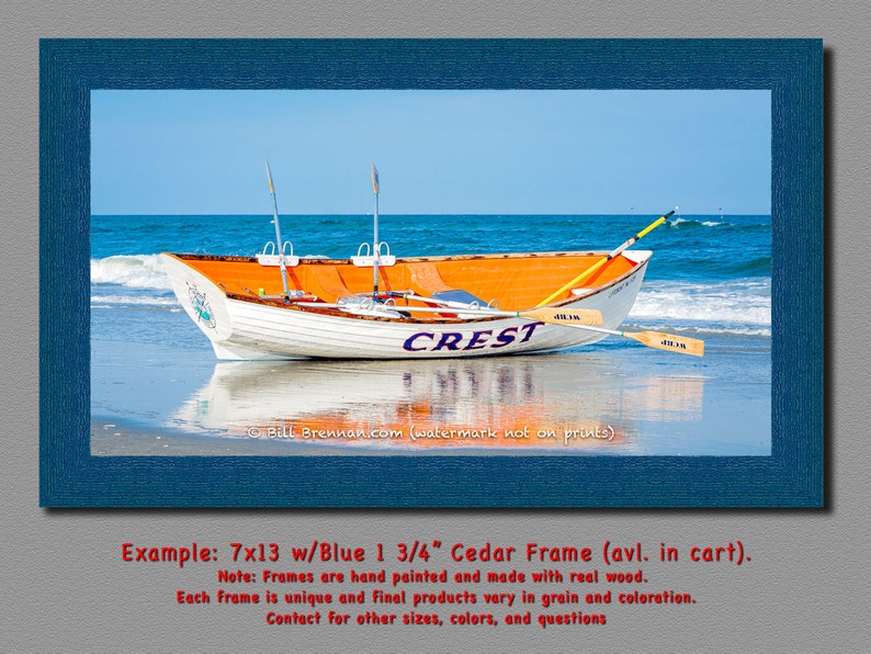May include: A framed print of a white and orange boat on a beach with the word "CREST" on the side. The boat has two oars and is set against a blue sky and ocean. The frame is blue and the print measures 7x13 inches.