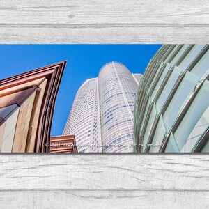 May include: Low-angle shot of modern skyscrapers against a clear blue sky. The buildings feature diverse architectural styles, including a tall, cylindrical tower and structures with glass facades and geometric designs. The image is a panoramic view.