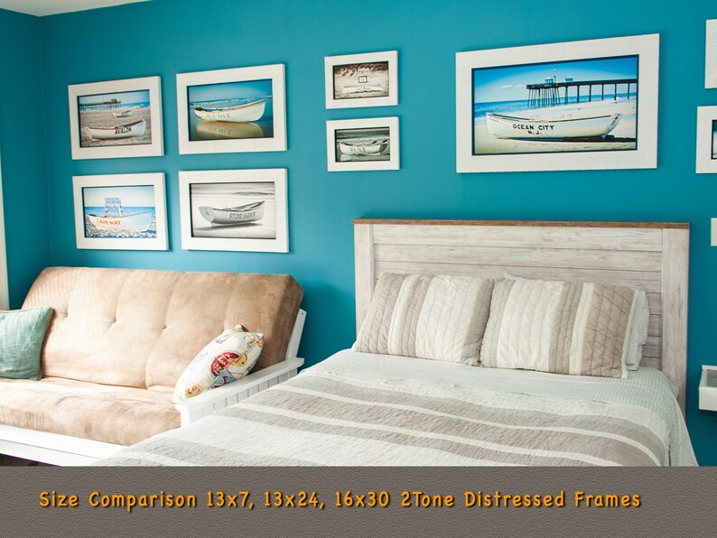 May include: A collection of framed beach-themed art prints in white distressed frames. The prints depict boats on sandy shores and a pier. The frames are displayed on a teal wall above a bed and futon. The text "Ocean City N.J." is visible on one print.