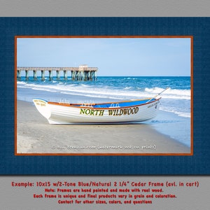 May include: A framed print showing a white boat on a sandy beach with the text "NORTH WILDWOOD". The boat has blue accents and is near the ocean with a pier in the background. The frame is blue with an orange border.