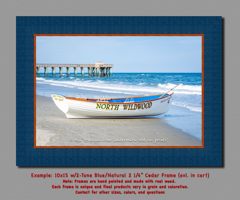 May include: A framed print showing a white boat on a sandy beach with the text "NORTH WILDWOOD". The boat has blue accents and is near the ocean with a pier in the background. The frame is blue with an orange border.