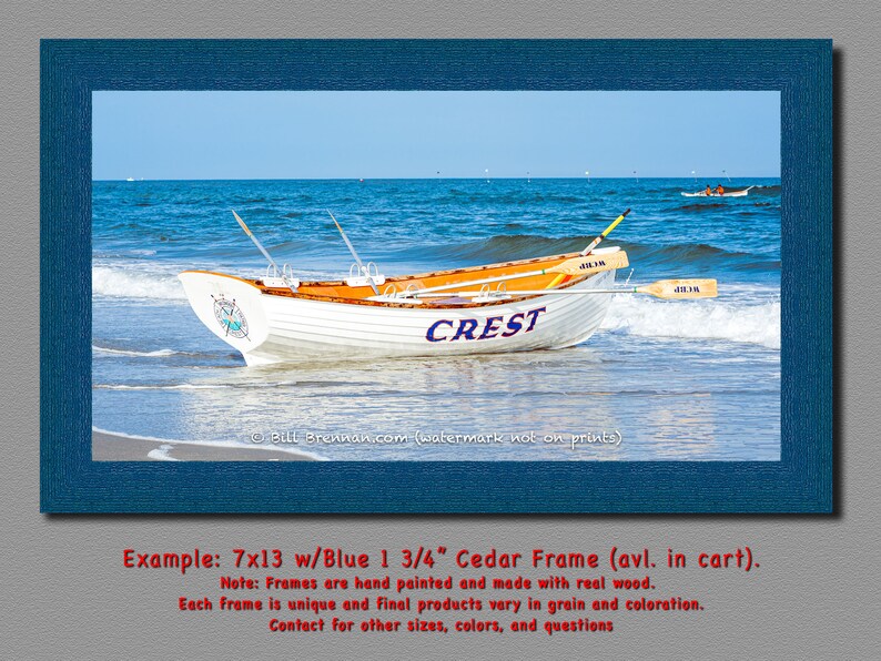 K&ouml;nnte beinhalten: Ein gerahmter Druck mit einem wei&szlig;en Boot mit dem Wort "CREST" an der Seite, das an einem Sandstrand liegt. Das Boot hat eine orangefarbene Innenausstattung und befindet sich in der N&auml;he von Meereswellen. Der Rahmen ist dunkelblau.