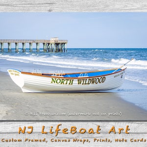 May include: A white and blue lifeboat on a sandy beach with the words "North Wildwood" printed on the side. The ocean and a wooden pier are in the background under a clear blue sky. The image is a print of NJ Lifeboat Art.