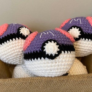 May include: Three crocheted Poke Balls in a brown burlap basket. The Poke Balls are purple, white, and black with pink accents.