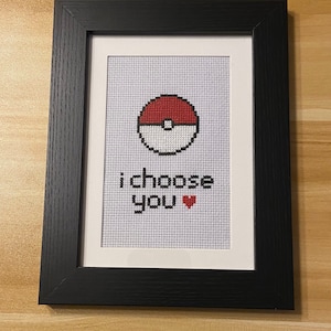 May include: A framed cross-stitch with a red and white Pokeball and the text "I choose you" with a red heart.