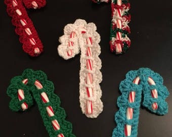 May include: Five crocheted candy cane ornaments in different colors. The ornaments are made with white, red, green, and blue yarn. The candy canes are wrapped in red and white striped paper.