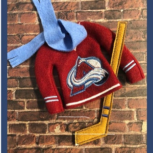 May include: A felt hockey jersey, scarf, and hockey stick set. The burgundy jersey features the Colorado Avalanche logo. The light blue scarf is draped over the jersey. A yellow hockey stick with blue stripes completes the set. The items are arranged on a brick background.