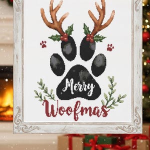 May include: Cross-stitch artwork in a white frame featuring a black paw print with the words "Merry Woofmas." The paw print is adorned with brown antlers, holly, and red paw prints. Green sprigs and red text complete the festive design.