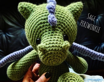 May include: Sage green crocheted dragon with a periwinkle colored horn and wings. The dragon is sitting on a person's hand.
