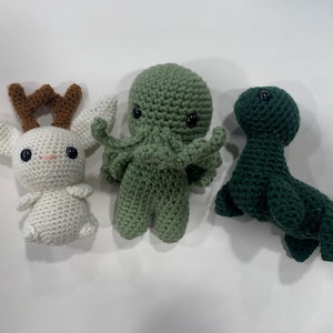 May include: Three handmade crochet toys: a white bunny with brown antlers, a green octopus-like creature, and a dark green seal. The toys are made with a textured stitch and have black safety eyes. The bunny has a pink nose.