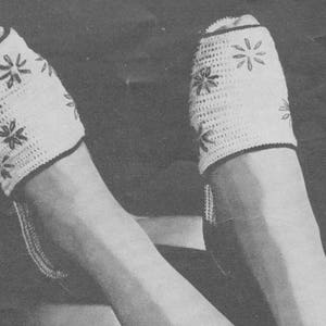 Op de afbeelding: Een paar witte gehaakte pantoffels met een bloemenpatroon. De pantoffels worden gedragen aan de voeten van een persoon.