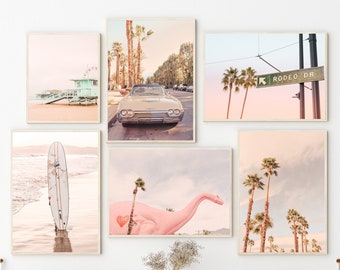 May include: A collection of framed art prints featuring coastal and California-themed scenes. The prints depict a lifeguard station, a classic car, a surfboard on the beach, a pink dinosaur, and palm trees. The color palette is soft, with pastel tones.