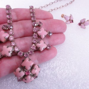 Op de afbeelding: Een roze strass ketting en oorbellen set. De ketting heeft een grote roze stenen hanger met kleinere roze stenen eromheen. De oorbellen zijn ook versierd met roze stenen.