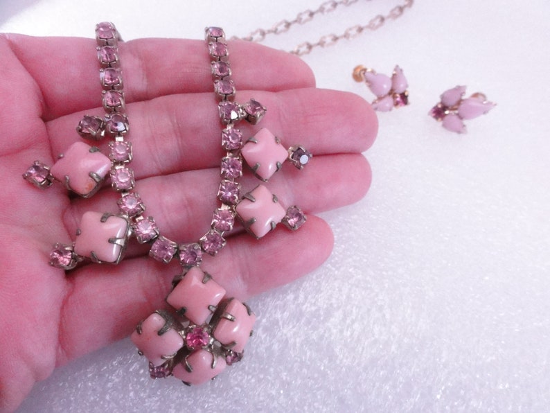 Op de afbeelding: Een roze strass ketting en oorbellen set. De ketting heeft een grote roze stenen hanger met kleinere roze stenen eromheen. De oorbellen zijn ook versierd met roze stenen.