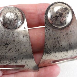 May include: Pair of vintage silver-toned metal earrings. Each earring features a rounded top and a triangular base with a rectangular cutout. The metal has a distressed, scratched appearance, suggesting age and wear. The earrings are likely for pierced ears.