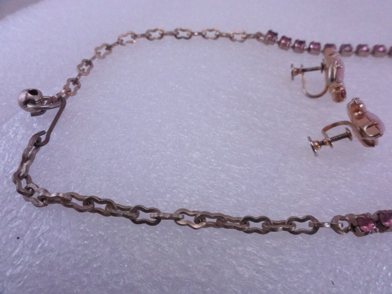 Op de afbeelding: Een zilveren ketting met een roze strass hanger en bijpassende oorbellen. De ketting heeft een sluiting en de oorbellen hebben schroefsluitingen.
