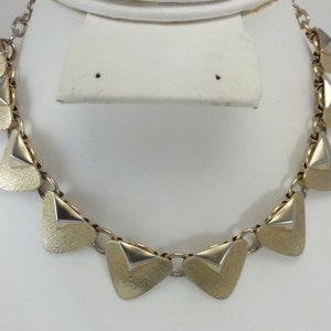 May include: A gold tone necklace with a chain and triangular pendant charms. The pendants are textured and have a silver tone accent.