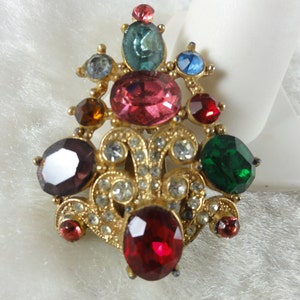 May include: A gold-tone brooch with a cluster of colorful rhinestones. The brooch features a large red rhinestone in the center, surrounded by smaller rhinestones in shades of pink, green, purple, blue, and brown.