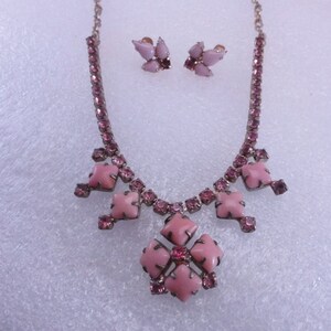 Op de afbeelding: Een roze strass ketting en oorbellen set. De ketting heeft een grote roze bloemvormige hanger met kleinere roze strass steentjes eromheen. De oorbellen zijn klein en hebben een roze bloem ontwerp.
