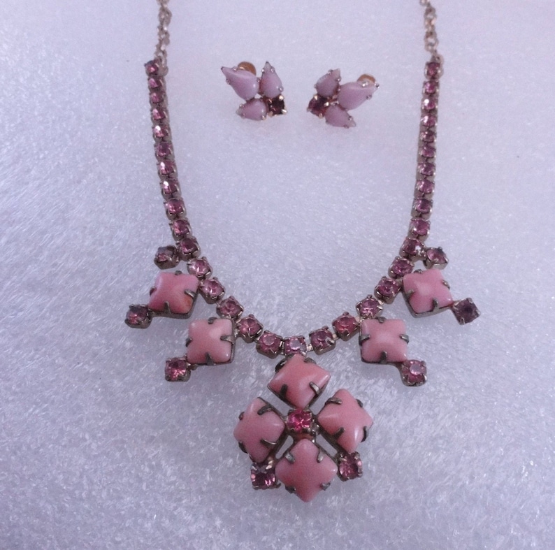 Op de afbeelding: Een roze strass ketting en oorbellen set. De ketting heeft een grote roze bloemvormige hanger met kleinere roze strass steentjes eromheen. De oorbellen zijn klein en hebben een roze bloem ontwerp.