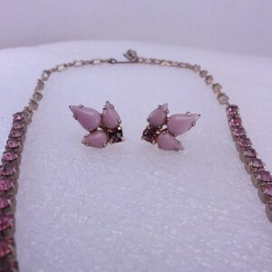 Op de afbeelding: Een roze strass ketting en oorbellen set. De ketting is een lange, dunne ketting met kleine roze strass steentjes. De oorbellen zijn een cluster van vijf roze strass steentjes.