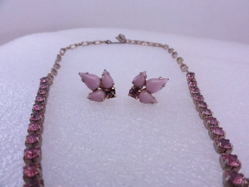 Op de afbeelding: Een roze strass ketting en oorbellen set. De ketting is een lange, dunne ketting met kleine roze strass steentjes. De oorbellen zijn een cluster van vijf roze strass steentjes.