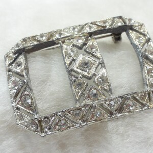 May include: A silver-toned metal buckle with a geometric design and clear rhinestones. The buckle has a rectangular shape with a smaller rectangular opening in the center.
