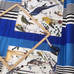 Puede incluir: Un marco de madera con un fondo de tela a rayas azul y blanco. El marco está colocado sobre una tela blanca con un patrón de pájaros coloridos. Un par de tijeras está sobre la tela.