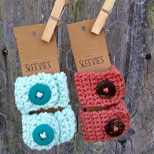 May include: Two crocheted cup sleeves, one teal and one coral, with wooden buttons. The sleeves are hanging on a wire fence with brown paper tags that say "Sleevies".