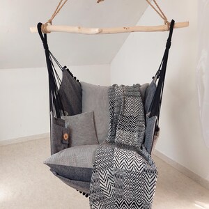 May include: A hanging chair featuring a light wood spreader bar and black rope suspension. The seat is upholstered in grey fabric with a small side pocket and a circular brown tag. It includes a grey cushion and a black and white chevron throw.