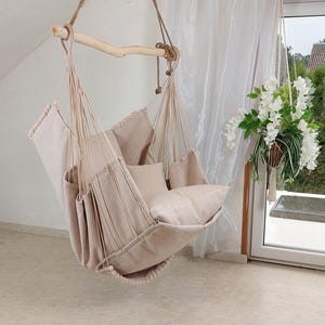 May include: A beige hanging chair made of fabric with pom-pom trim, suspended by thick ropes from a natural wooden bar. The chair includes a seat cushion and a back cushion, featuring a textured weave pattern throughout the entire structure.