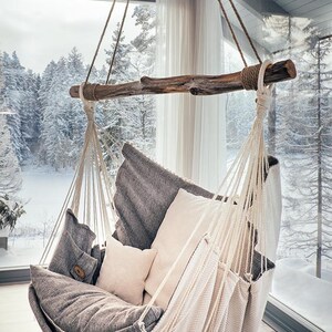 May include: A hanging chair suspended from a thick, natural wooden branch by cream-colored ropes. The seat features a dark gray fabric base, a matching gray cushion, and two smaller cream-colored square pillows, all supported by a woven fabric frame.