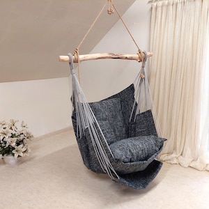 Puede incluir: Una silla colgante con asiento y respaldo de tela texturizada gris oscuro, suspendida mediante cuerdas de color gris claro de una rama de madera natural de tono claro. La estructura se sostiene con un lazo de cuerda grueso en el centro.