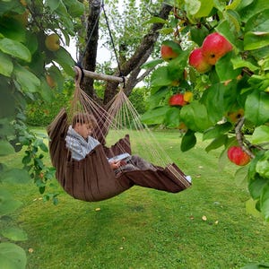 Puede incluir: Una silla colgante de tela marrón suspendida por cuerdas gruesas de una barra separadora de madera. La silla presenta un material texturizado y está sujeta a una rama de árbol. El asiento está diseñado para descansar al aire libre en un jardín.