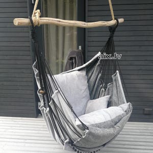 May include: A hanging chair featuring a light gray fabric seat with a textured weave, suspended by black ropes from a natural wooden branch. The chair includes two matching gray cushions and is accented with small black pom-pom trim along the edge.